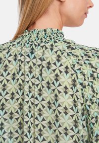 Musterbluse mit gerafftem Ausschnitt. Sie hat ein sich wiederholendes geometrisches Blumenmuster in Hellgrün und sanftem Blau auf einem dunklen Hintergrund.