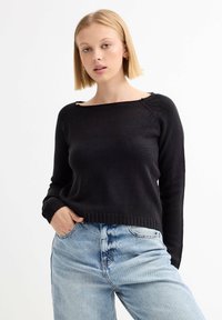 Suéter negro de punto con un escote cuadrado y puños acanalados, combinado con jeans de talle alto en azul claro, mostrando un ajuste casual.