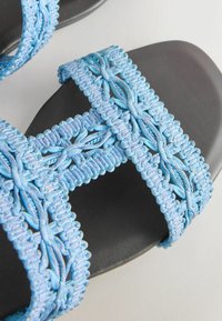 Sandales plates noires avec de larges brides tressées bleu clair traversant les orteils et la voûte plantaire.