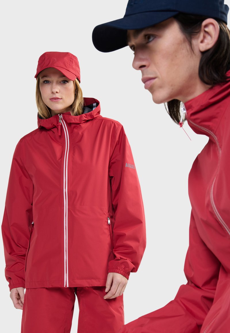 Rode waterdichte jas met ritssluiting, elastische manchetten en een capuchon. Gladde textuur, gecombineerd met bijpassende broek. Marineblauwe pet gedragen door het model.