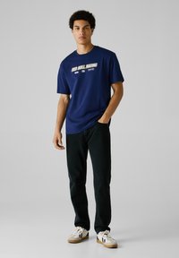 Młody mężczyzna stoi z ręką w kieszeni, ubrany w granatowy T-shirt Red Bull Racing, czarne dżinsy i białe sneakersy na jednolitym jasnym tle.