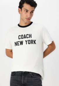 White cotton t-shirt with black ringer collar, featuring bold black text: "COACH NEW YORK" centered across the chest. Casual fit.
