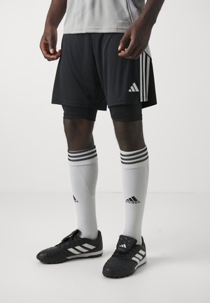 Man draagt zwarte Adidas sportshorts, witte Adidas kniekousen met zwarte strepen en zwarte Adidas voetbalschoenen op een witte achtergrond.