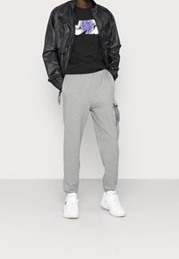 Schwarze Zip-Up-Jacke, schwarzes T-Shirt mit "JUMP MAN" Grafik, graue Jogginghose mit einer seitlichen Cargo-Tasche und weiße Sportschuhe.