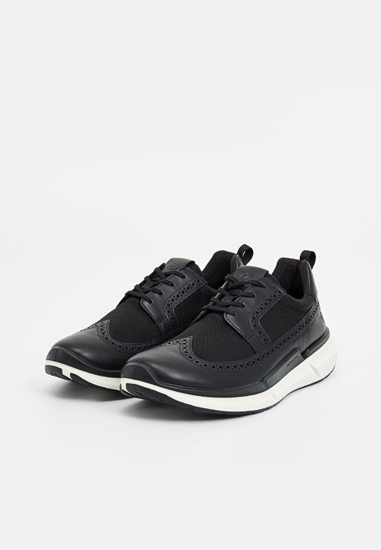 Une paire de baskets noires en cuir et en mesh, à lacets, avec des semelles blanches, des détails en ailes et des languettes, sur fond blanc.