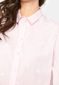 Ljusrosa skjorta med krage och knappstängning, prydd med vita broderade palmer och en texturerad tygkvalitet.