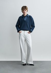 Jeune personne portant une chemise à manches longues bleu marine avec col, un pantalon large blanc, et des chaussures pointues noires, debout devant un fond uni.