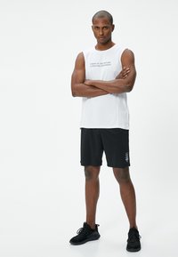 Top atlético blanco sin mangas con texto impreso, combinado con pantalones cortos negros con un sutil patrón geométrico y zapatillas deportivas negras.