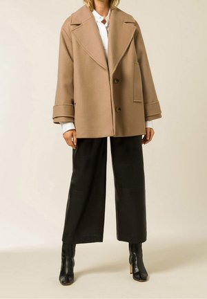 Modèle portant un manteau croisé couleur camel, une chemise blanche, un pantalon noir à jambes larges et des bottines noires, devant un fond de studio uni.