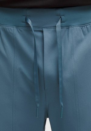 Mėlynos sportinės kelnės iš arti, matomas raištelių juosmuo ir smulkios vertikalios siūlės audinyje.