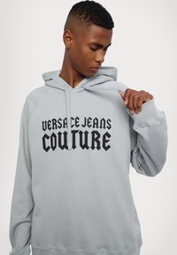 Ανοιχτό γκρι φούτερ με κουκούλα και μαύρο λογότυπο "VERSACE JEANS COUTURE". Διαθέτει κουκούλα με κορδόνι και ραμμένες μανσέτες για επιπλέον υφή.