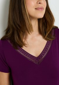 T-shirt viola a scollo a V con maniche corte, caratterizzato da una delicata rifinitura in pizzo lungo il colletto. Tessuto morbido con una consistenza liscia e una leggera lucentezza.