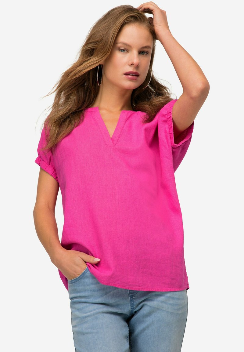 Blusa corta in lino rosa con maniche a giro e scollatura a V, caratterizzata da una vestibilità ampia e una texture morbida. Indossata con jeans azzurri.