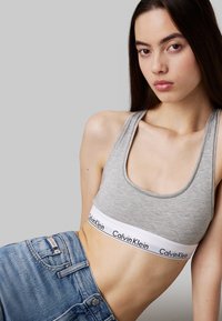 Sujetador deportivo gris de algodón con banda blanca ancha que presenta el logo "Calvin Klein"; combinado con jeans de mezclilla azules. Textura suave, diseño casual.