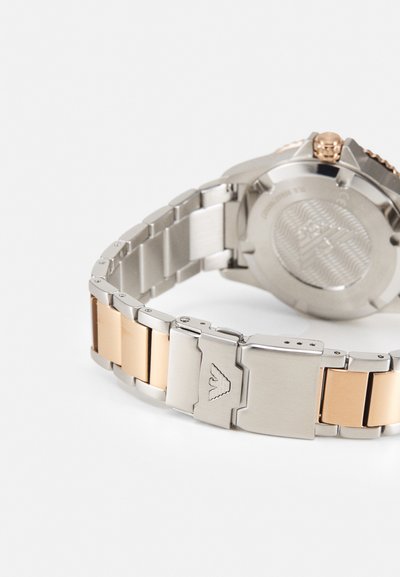 Silberne und goldfarbene Metalluhr mit einem Metallgliedarmband; verfügt über eine geb brushed Oberfläche; beinhaltet einen Verschluss mit Markenlogo an der Seite.