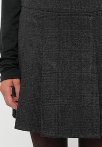 Grå skjortsbar med folder laget av teksturert stoff med subtile mønstre; stylet med en svart langermet topp delvis synlig.