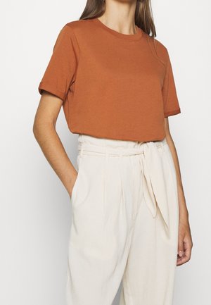 Kobieta w rdzawej, krótkim rękawie, bluzce z okrągłym dekoltem, wsuniętej w kremowe, wysokokatne spodnie typu paperbag z przewiązanym paskiem. Jedna ręka w kieszeni.