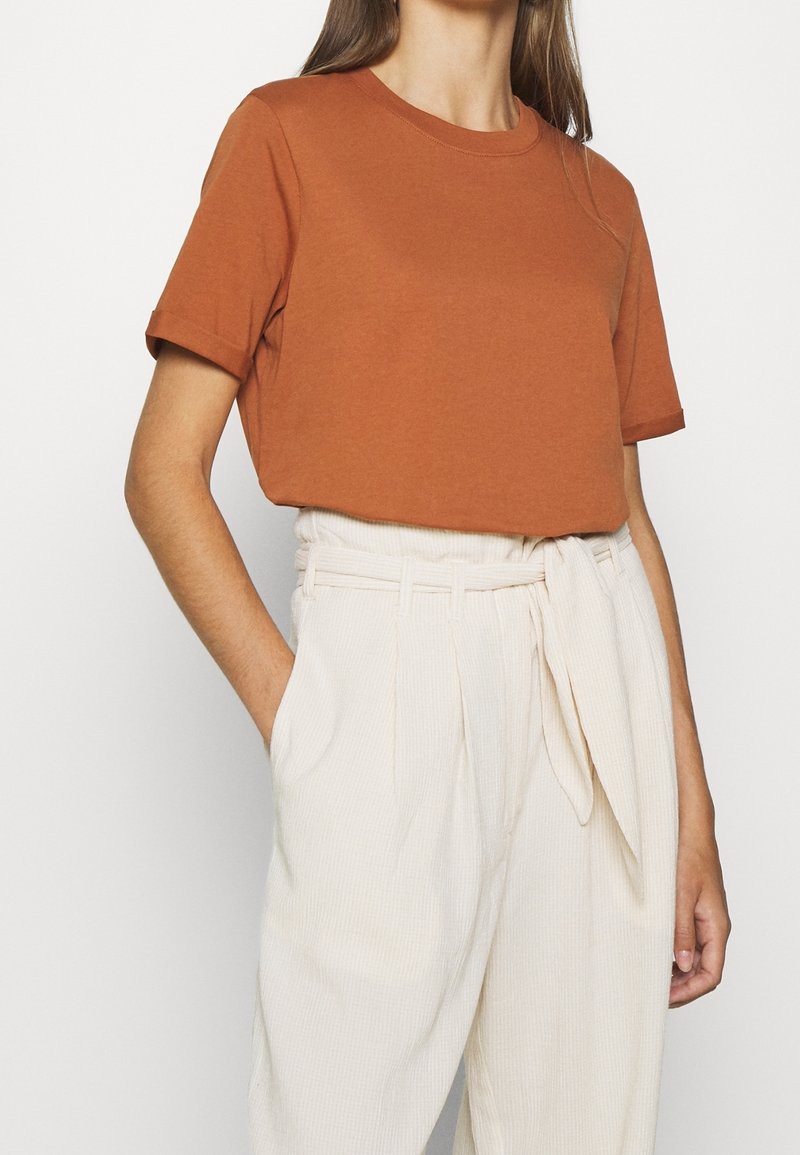 Donna che indossa una maglietta a maniche corte color ruggine con scollo rotondo infilata in pantaloni a vita alta color crema con cintura annodata in stile paperbag. Una mano in tasca.