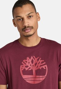 Marinbomullströja med en stor röd trädlogotyp på framsidan, rund halsringning och korta ärmar. Slät textur med en klassisk passform.