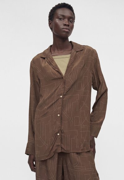 Μαύρη γυναίκα που φοράει καφέ πουκάμισο με σχέδια και κουμπιά και ταιριαστό παντελόνι πάνω από μια μπεζ μπλούζα, στέκεται με το χέρι στην τσέπη της.