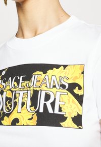 Vit bomullströja med en svart rektangulär grafik med gula utsmyckningar och fet vit text som står "VERSACE JEANS COUTURE."