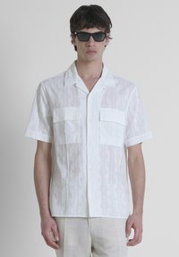 Camicia bianca a maniche corte con un motivo geometrico, dotata di due tasche sul petto e chiusura con bottoni. Tessuto liscio e leggero.