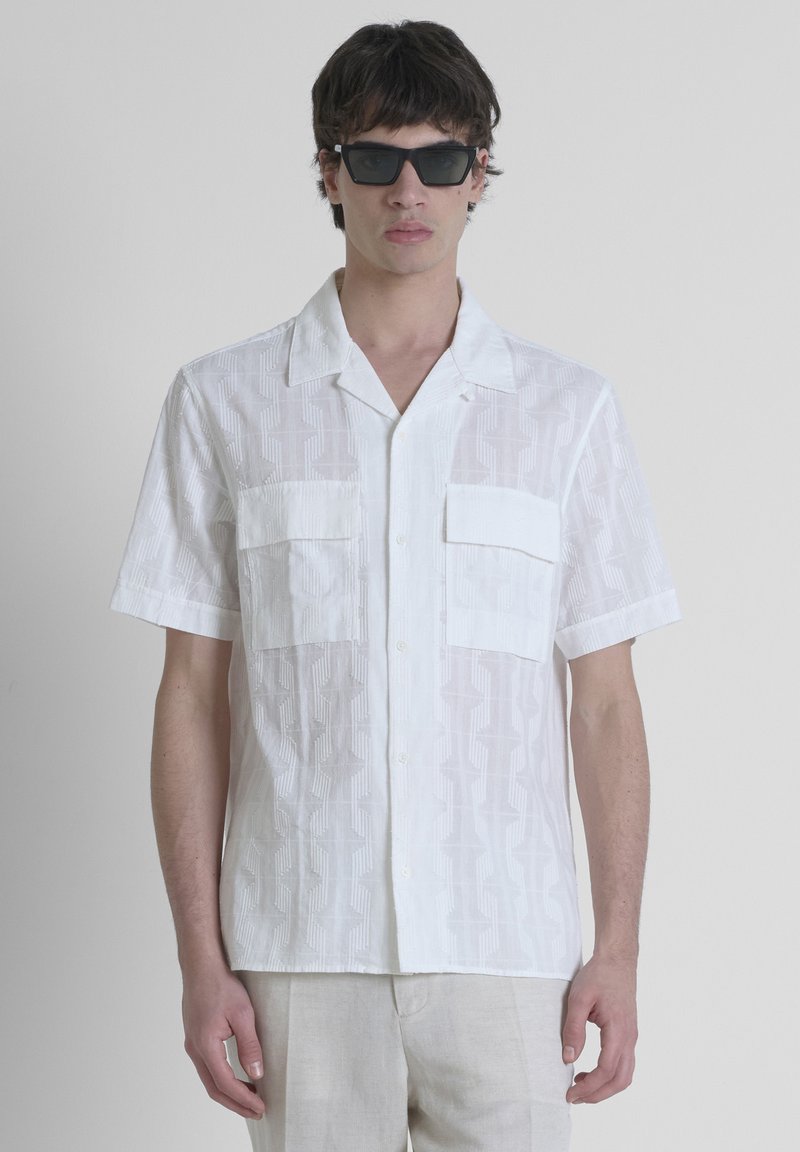 Camicia bianca a maniche corte con un motivo geometrico, dotata di due tasche sul petto e chiusura con bottoni. Tessuto liscio e leggero.