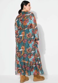 Langes, blumiges Kleid mit einem türkisfarbenen Hintergrund und orangefarbenen sowie rosafarbenen Blumen. Fließendes Design mit Rüschensaum und transparenter Textur. Mit tanfarbenen Stiefeln kombiniert.