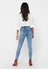 Kvinna som står med ryggen vänd, klädd i ljusblå skinny jeans, en vit blus med puffärmar och bruna mocka ankellånga stövlar med klackar.