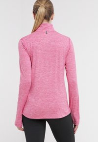 Frau mit blonden Pferdeschwanz, die ein langärmliges, rosa meliertes Oberteil und schwarze Leggings trägt, mit dem Rücken zu einer einfarbigen weißen Hintergrund.