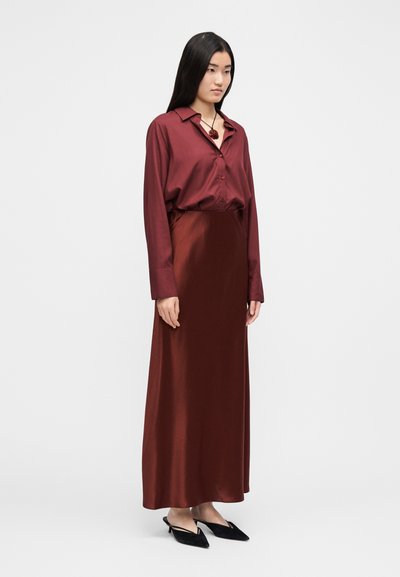Bordowa satynowa bluzka z długimi rękawami i kołnierzykiem, zestawiona z pasującą długą spódnicą. Całość dopełniają czarne buty z szpiczastym noskiem.