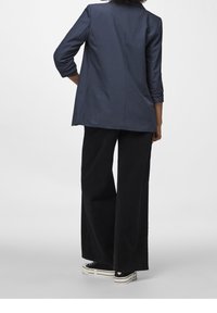 Personne portant un blazer bleu marine à manches retroussées, un pantalon noir à jambes larges et des baskets compensées noires, debout devant un fond uni.