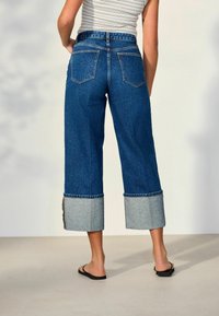 Persona vistiendo vaqueros azules de pierna ancha con grandes puños, una camiseta sin mangas a rayas y chanclas negras, de pie sobre un suelo claro.