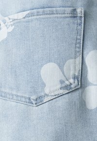 Lys blå denimstoff med et hvitt blomsterdesign. Inkluderer en lapp lomme med sydd kontur og synlig tekstur.