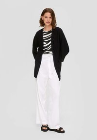 Schwarze offene Strickjacke über einem gestreiften Schwarz-Weiß-Oberteil, kombiniert mit lockeren weißen Hosen und schwarzen Sandalen, vor einem grauen Hintergrund präsentiert.