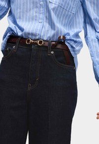 Ingrandimento di una persona che indossa jeans blu scuri e una camicia a righe blu infilata in una cintura di pelle marrone con una fibbia dorata.