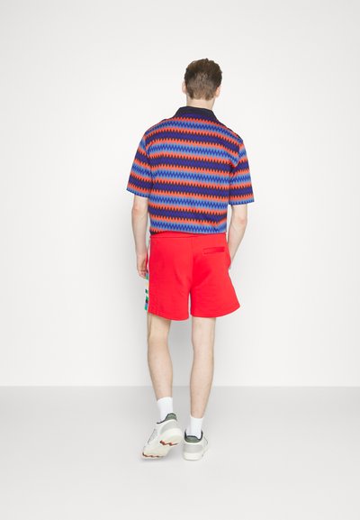 Chemise à manches courtes avec un motif en zigzag bleu, rouge et noir, assortie à un short rouge uni et des baskets blanches avec des accents verts.