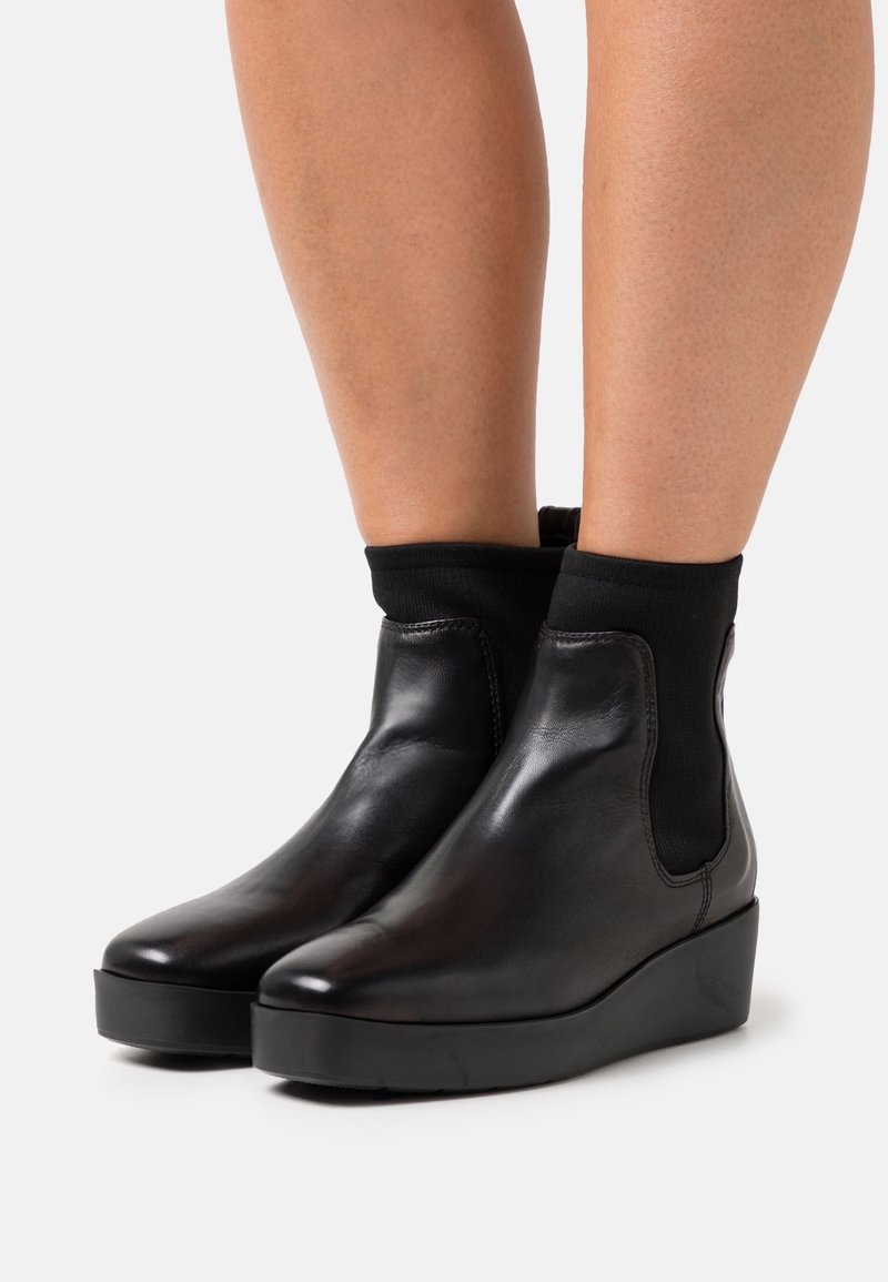 Bottes compensées en cuir noir montant jusqu'à la cheville, portées avec des chaussettes noires sur des jambes nues, sur fond blanc.