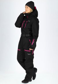 Black waterproof ski suit with pink accents, zip pockets, and a belt. Includes a hood and elastic wrists, designed for outdoor activities.