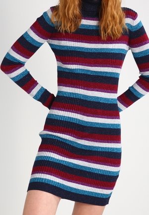 Vrouw in een aansluitende, gebreide jurk met lange mouwen en horizontale strepen in marineblauw, bordeaux, blauw en wit.