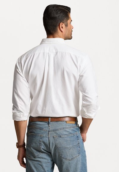 Chemise blanche à manches longues avec un col boutonné, associée à un jean en denim bleu. La chemise est montrée de dos, avec les manches retroussées.