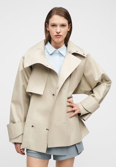 Γυναίκα που φοράει μια μπεζ υπερμεγέθη καμπαρντίνα πάνω από ένα ανοιχτό γαλάζιο πουκάμισο και γκρι σορτς, στέκεται με το ένα χέρι στη μέση.