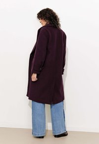 Long manteau violet en tissu épais, avec un col cranté, des poches latérales et des poignets boutonnés. Assorti à un jean bleu à jambes larges.