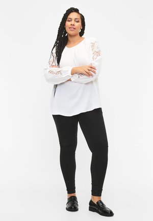 Vit blus med spetsdetaljer på ärmarna, kombinerad med svarta leggings och svarta snörskor. Slät tyg, avslappnad passform, långa ärmar.