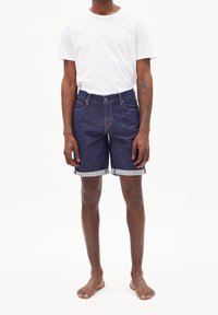 Shorts in denim blu navy con orli arrotolati, tasche frontali e chiusura con bottone, abbinati a una maglietta bianca semplice. Il tessuto è morbido e liscio.