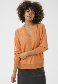 O orange pulover s V-izrezom in ohlapnim krojem, z rebrastimi manšetami in robom; mehak material, nosi se čez črno usnjeno krilo.