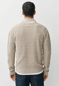 Man met kort, krullend haar, gekleed in een beige gebreid vest en donkerblauwe jeans, gezien van achteren tegen een witte achtergrond.
