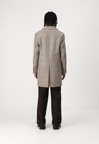 Abrigo de cuadros marrón con un corte entallado, solapas de picos y puños abotonados. Combinado con pantalones oscuros, que muestran una textura suave y líneas limpias.