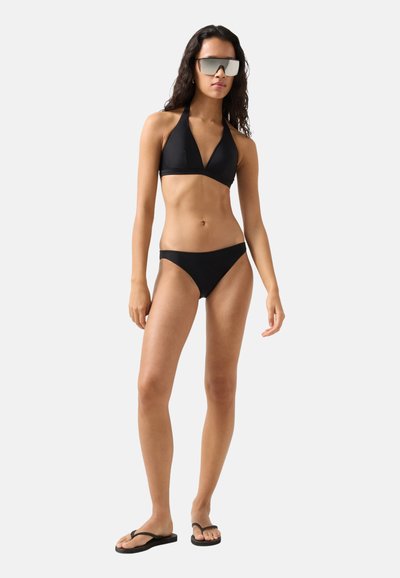 Kvinde iført sort halter-bikini, sorte flip-flops og store reflekterende solbriller, stående foran en ensartet lys grå baggrund.