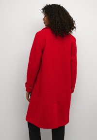 Rode, gestructureerde jas met een kraag en knoopmanchetten, met een losse pasvorm en een rugsplit voor gemakkelijke beweging. Zwarte broek zichtbaar daaronder.